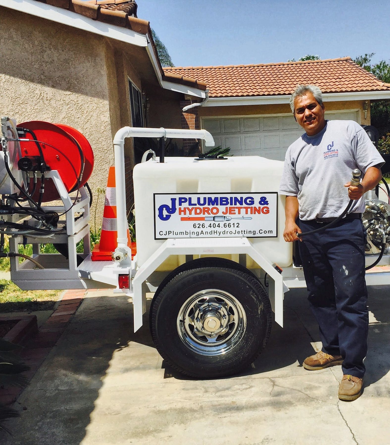 Carlos, the owner of CJ Plumbing & Hydro Jetting, smiling beside his white hydro-jetting trailer with the CJ sign and red hose reel